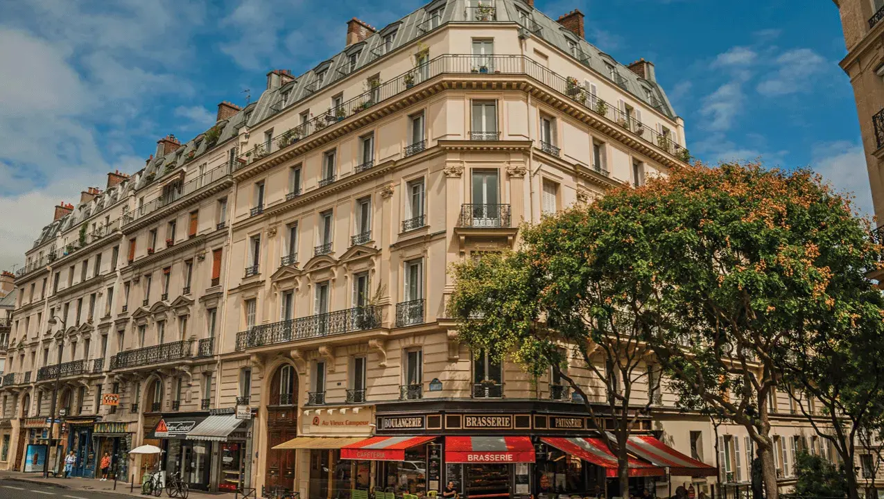 Immeuble parisien typique de l'architecture haussmannienne, avec ses balcons en fer forgé et ses toits mansardés. Un arbre fleuri borde la façade.