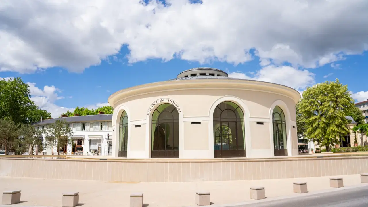 Le pavillon de la Source de l'Hôpital à Vichy, un lieu chargé d'histoire, se dresse sous un ciel bleu et nuageux.