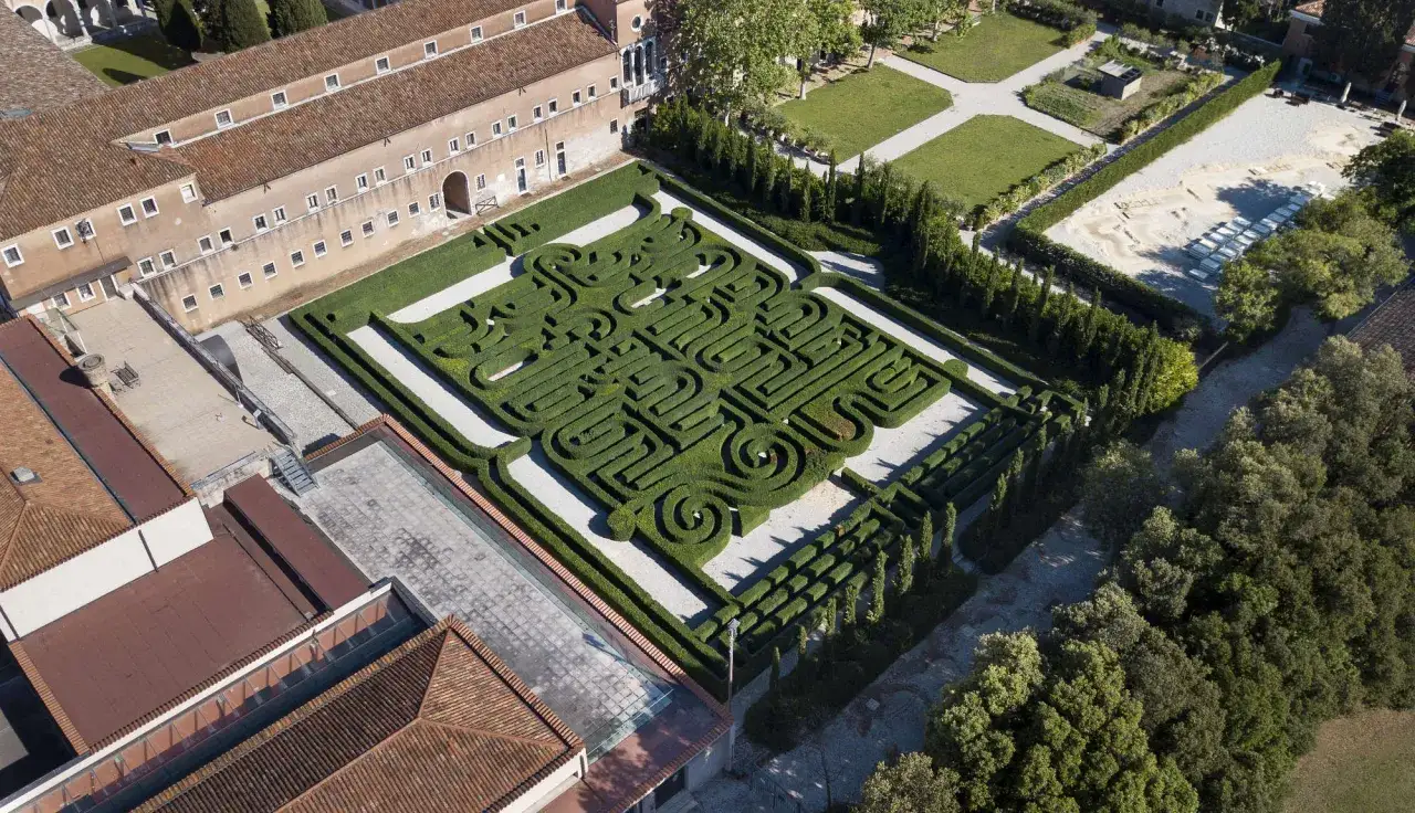 Vue a&eacute;rienne d'un labyrinthe de haies taill&eacute;es, &eacute;voquant une biblioth&egrave;que de Babel verdoyante, entour&eacute; de b&acirc;timents aux toits de tuiles rouges.